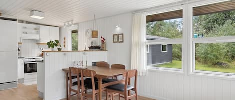 A modern kitchen with white cabinetry, a dining area with wooden chairs, and large windows offering a view of the outdoors.
