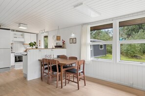 A modern kitchen with white cabinetry, a dining area with wooden chairs, and large windows offering a view of the outdoors. - Cozy holiday home in Hune near Blokhus (Hune)