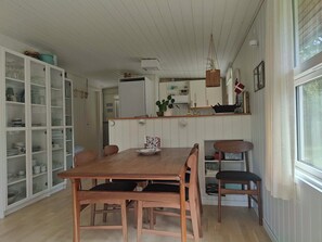 A dining area with a wooden table and chairs, a kitchen with white cabinets, and a window with curtains. - Cozy holiday home in Hune near Blokhus (Hune)