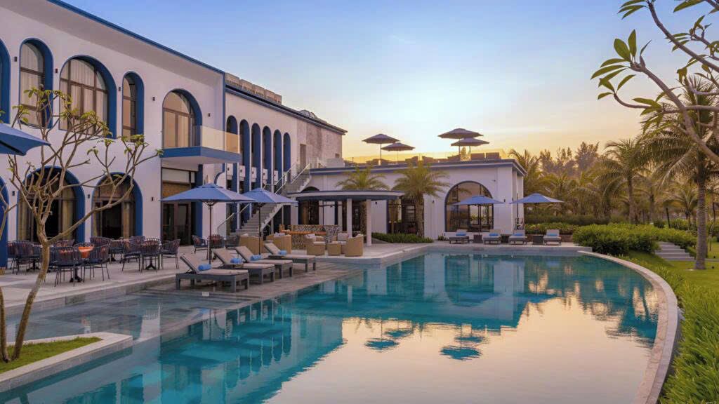 Un espace piscine avec des chaises longues et des parasols, un bâtiment doté de fenêtres en arc de cercle et un ciel dégagé.