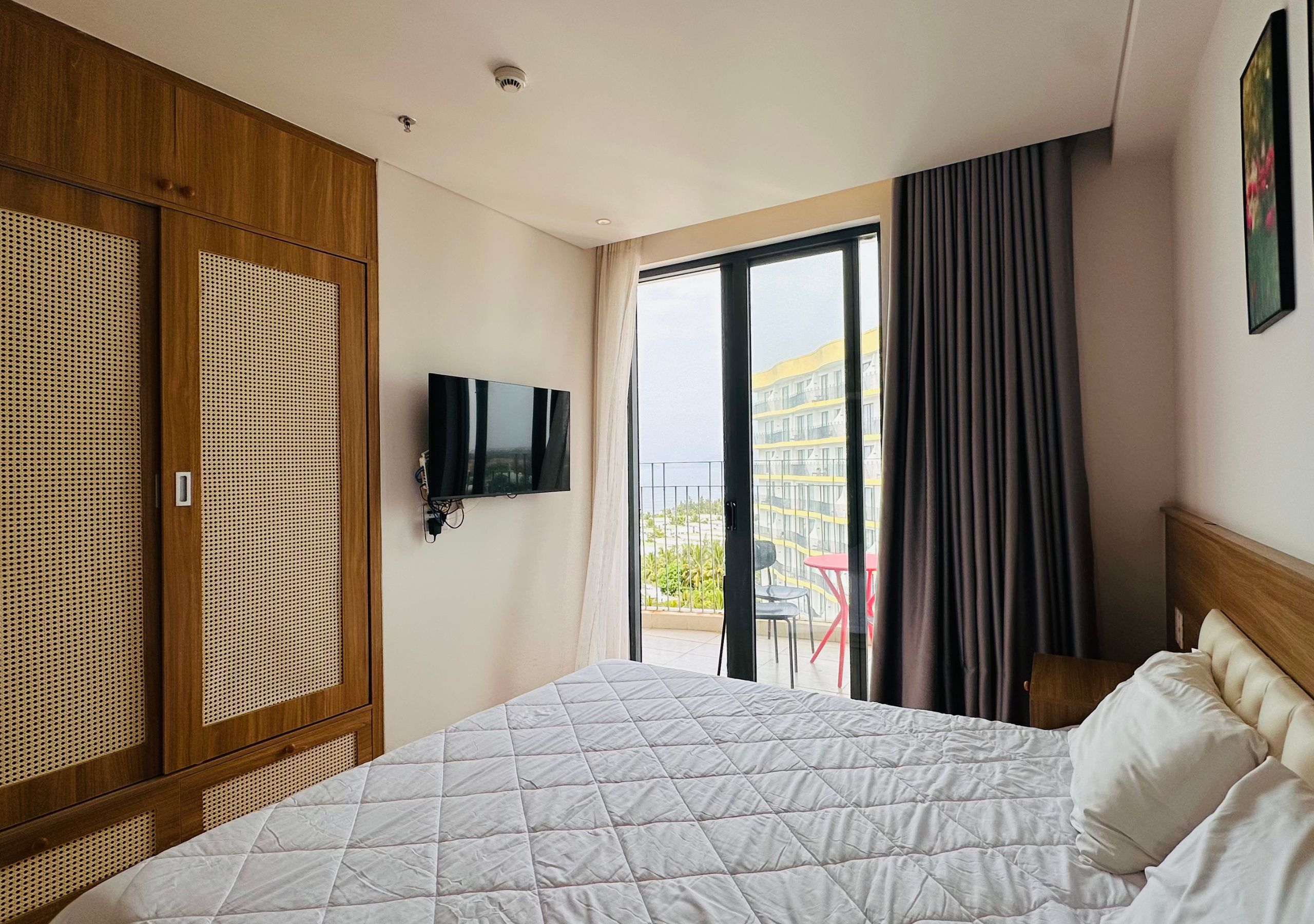 Habitación de hotel con cama, televisor, puerta corrediza de vidrio a un balcón y armario de madera.