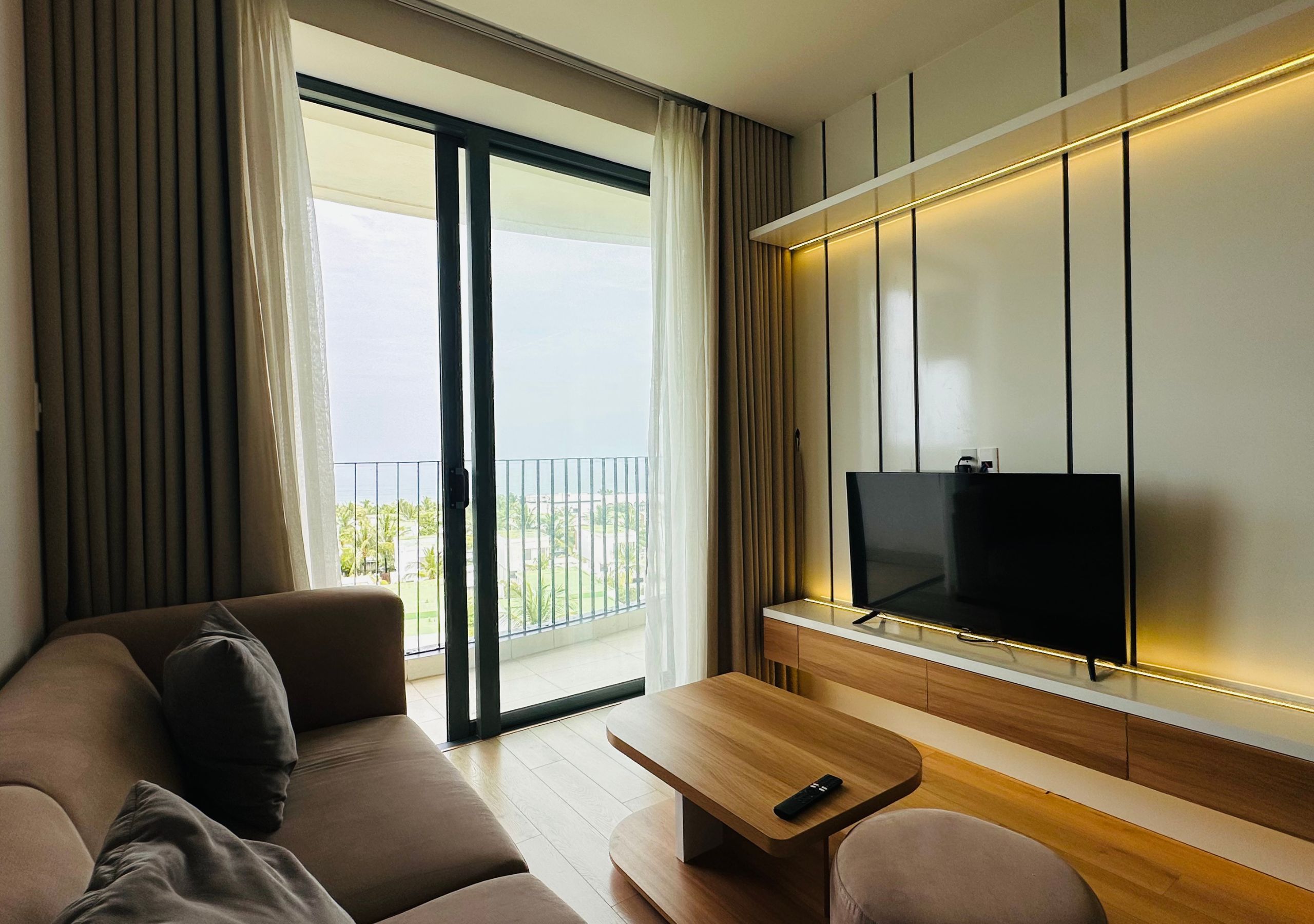 A modern hotel room with a sofa, a coffee table, a TV, and a balcony with curtains.