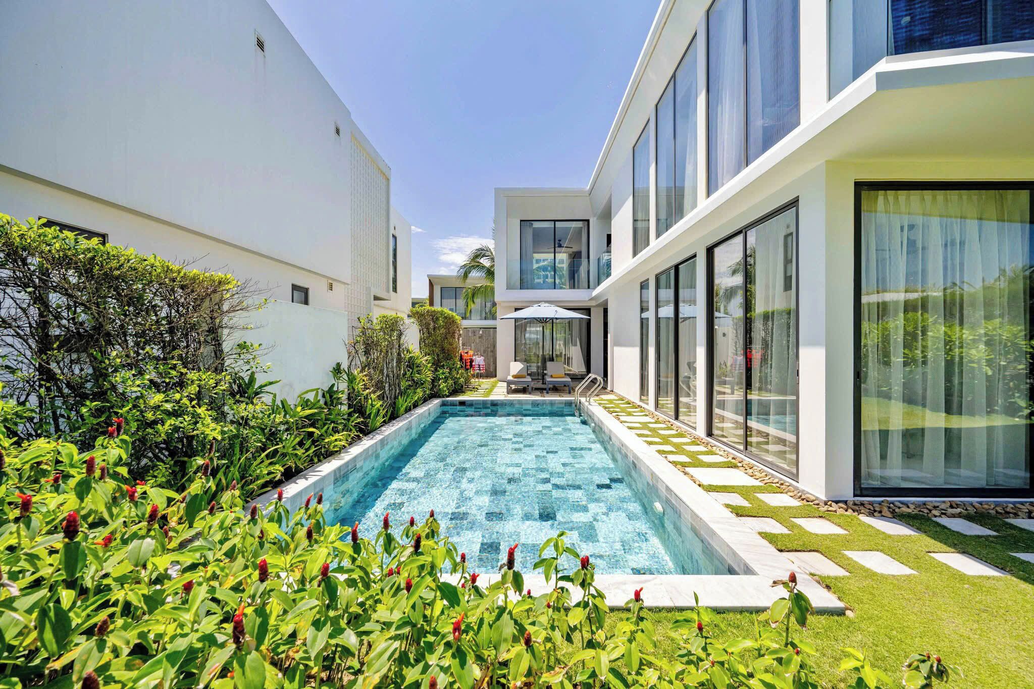 Un área de piscina moderna con una piscina azul cristalina, rodeada de exuberante vegetación y un edificio blanco con amplias ventanas.