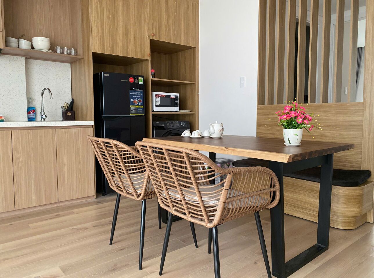 A modern kitchen with wooden cabinets, a black refrigerator, a microwave, and a dining table with wicker chairs.