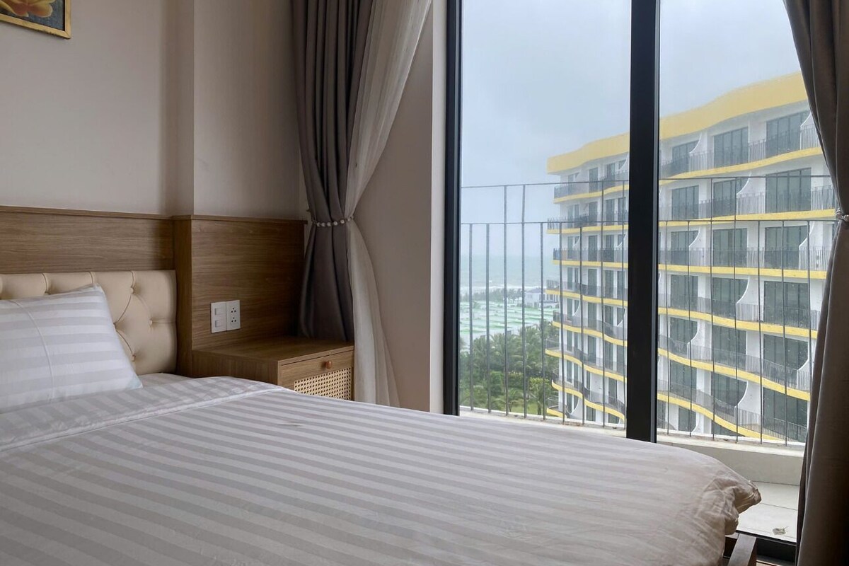 A hotel room with a large bed, a bedside table, a lamp, and a view of a high-rise building.