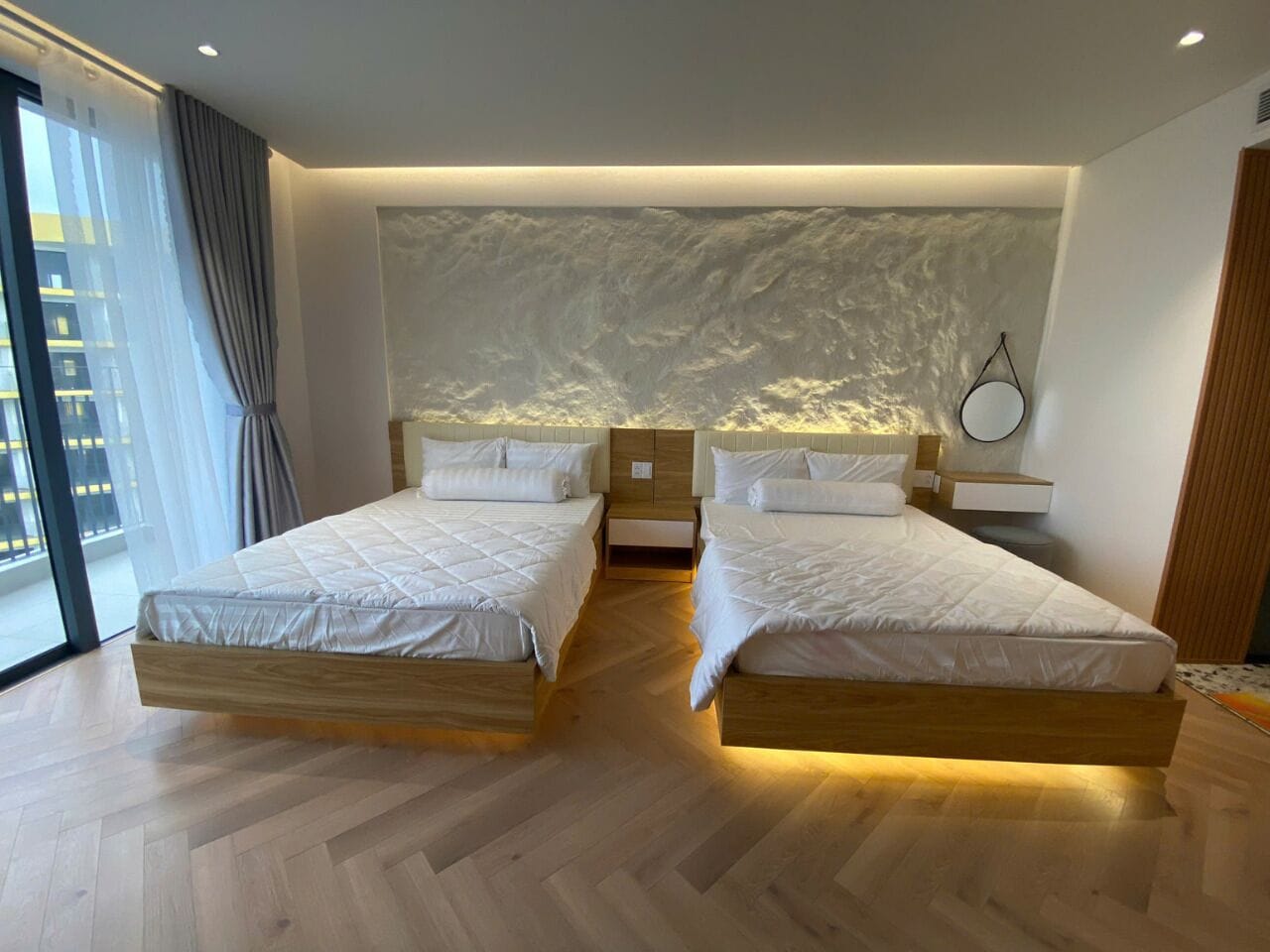 A hotel room with two beds, a large window with curtains, a wall-mounted artwork, and a wooden floor.