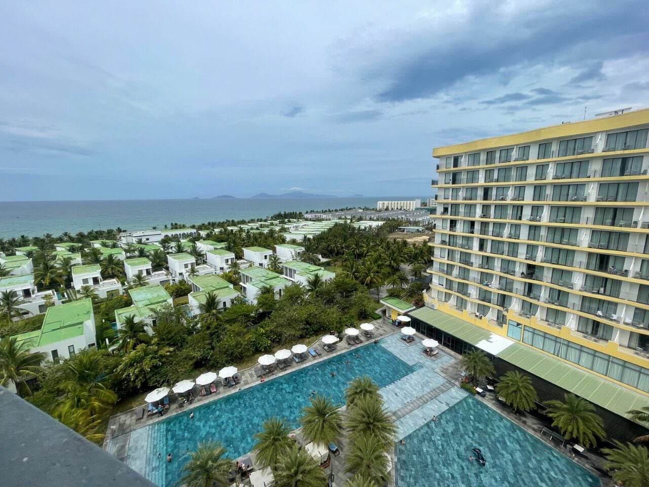 Hotel com uma piscina grande, cercada por palmeiras e vista para o mar.