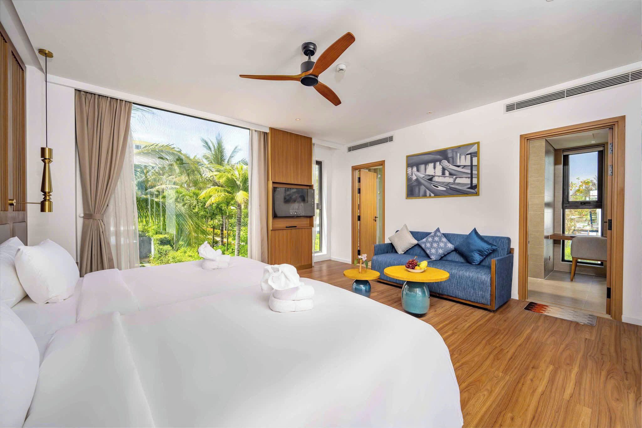 Une chambre d’hôtel avec un grand lit, un ventilateur de plafond, une télévision, un canapé avec des coussins et une vue sur des palmiers.