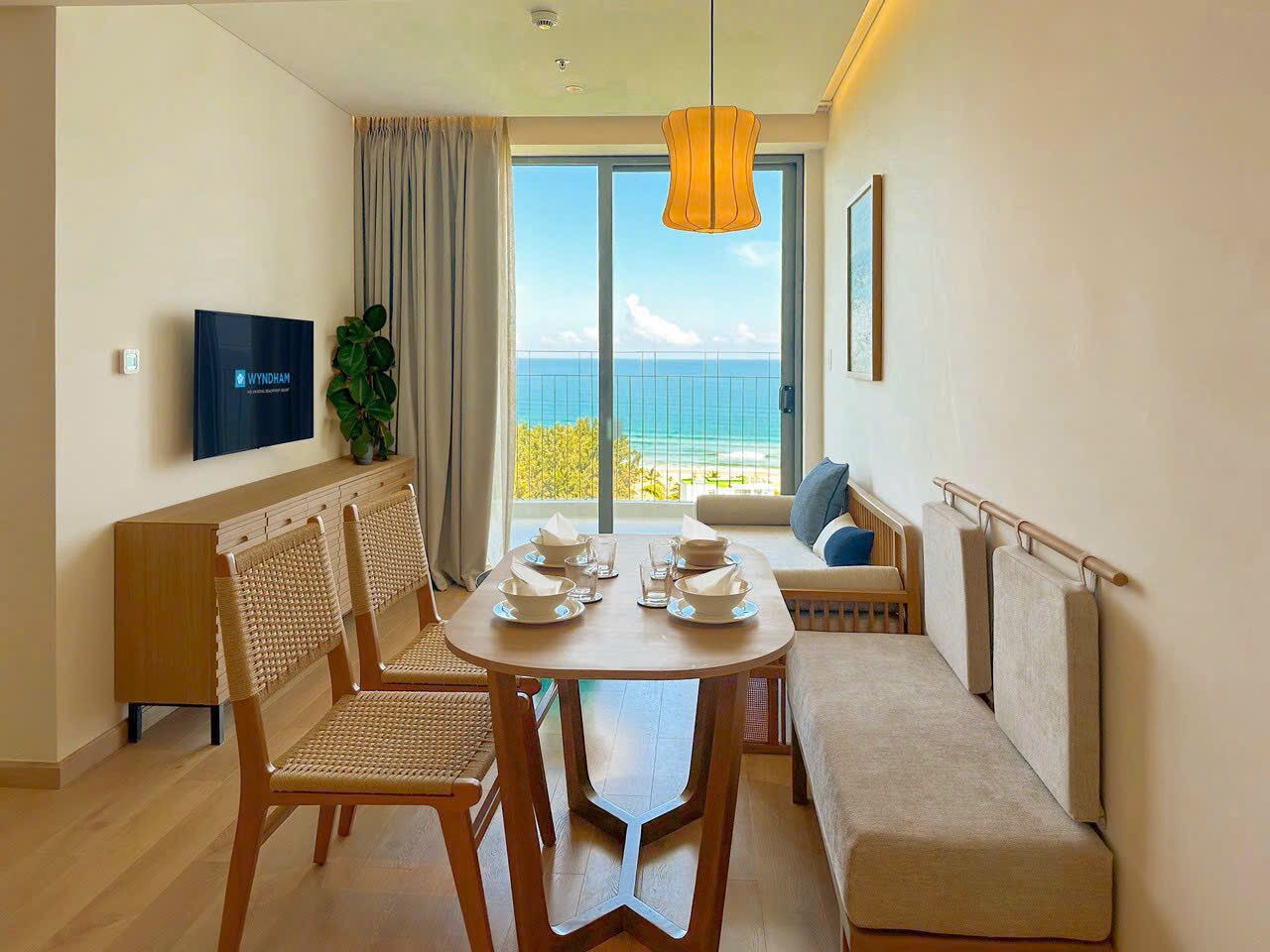 A hotel room with a dining table set for four, a sofa, a TV mounted on the wall, and a large window with a view of the sea.