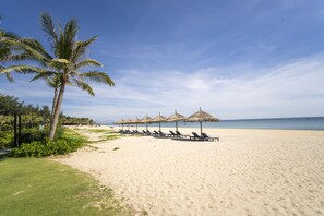 A beach with sun loungers, palm trees, and thatched umbrellas.