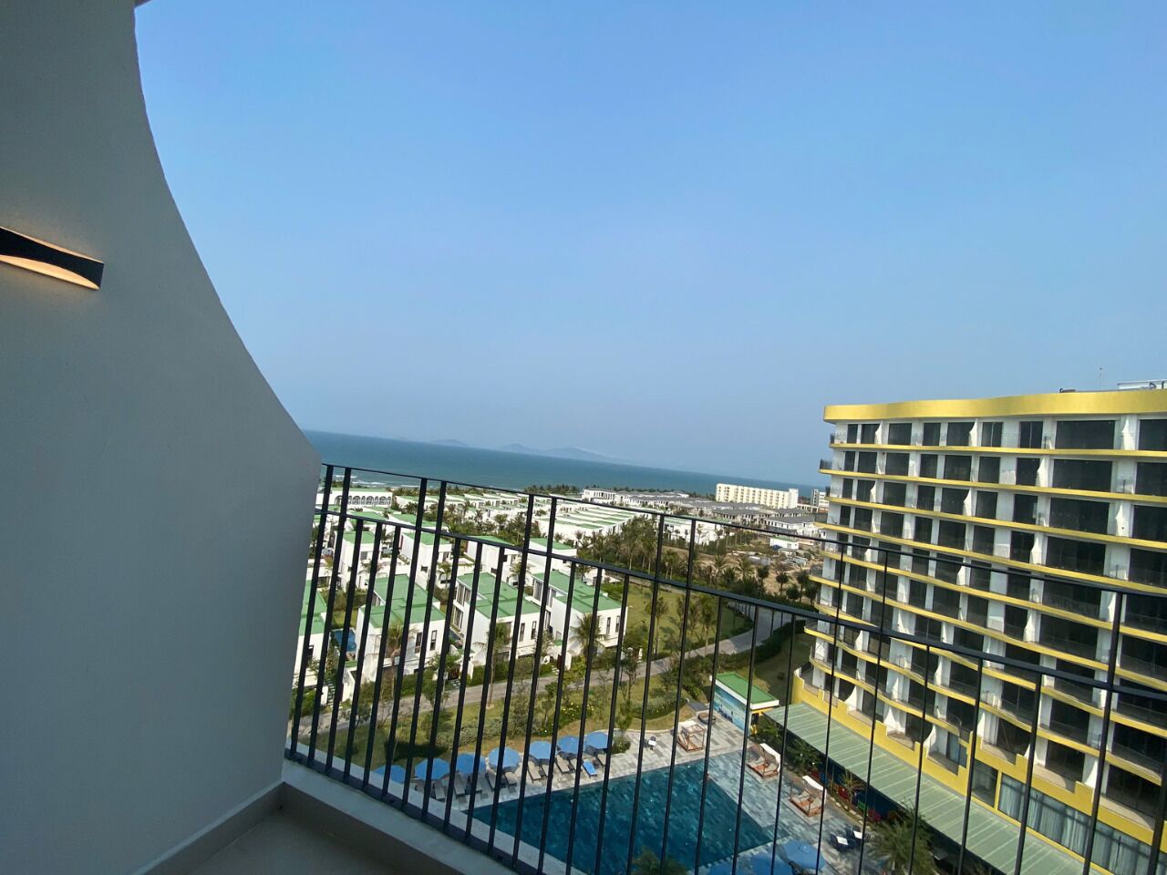 Un balcón con vistas a una piscina, un edificio de hotel y una costa lejana.