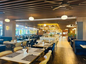 Een ruime eetzaal met hoge plafonds, houten lambrisering en een bar in het midden. Tafels staan netjes klaar met servies.