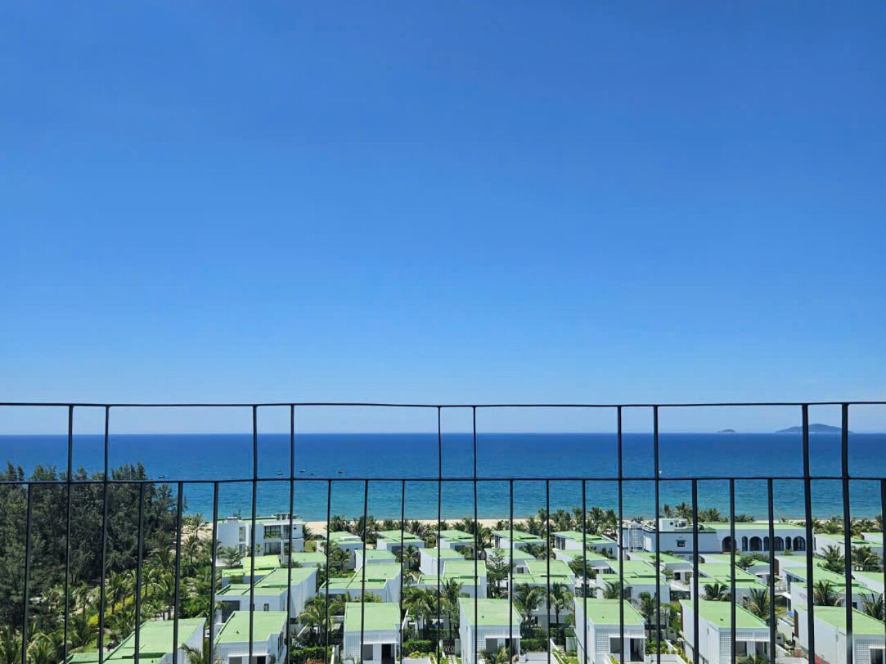 Vista costera con cielo despejado, balcón con barandilla metálica y varios edificios con tejados verdes.