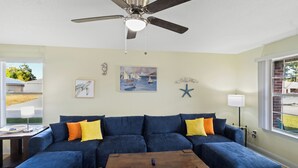 A living room with a blue sectional sofa, a wooden coffee table, a ceiling fan, and a window with a view of the outdoors. - Sunshine & Shenanigans | Navarre | 5 Min to Beach (Navarre)