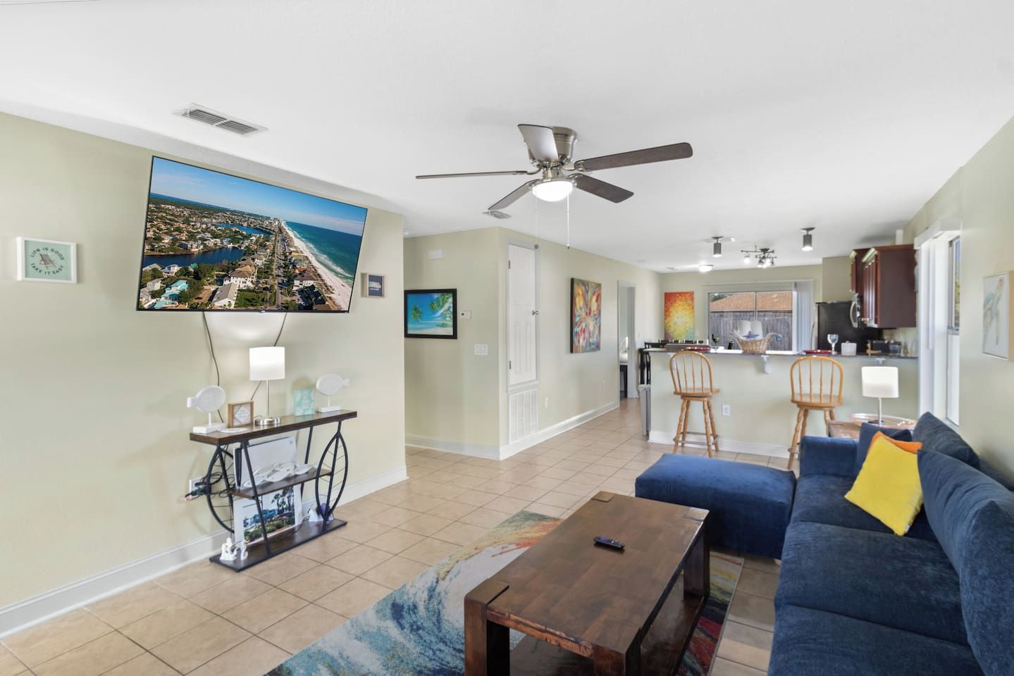 A spacious living area with a large TV, a ceiling fan, a kitchen in the background, and a blue sofa.