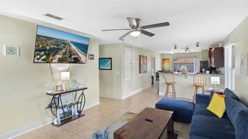 A spacious living area with a large TV, a ceiling fan, a kitchen in the background, and a blue sofa.