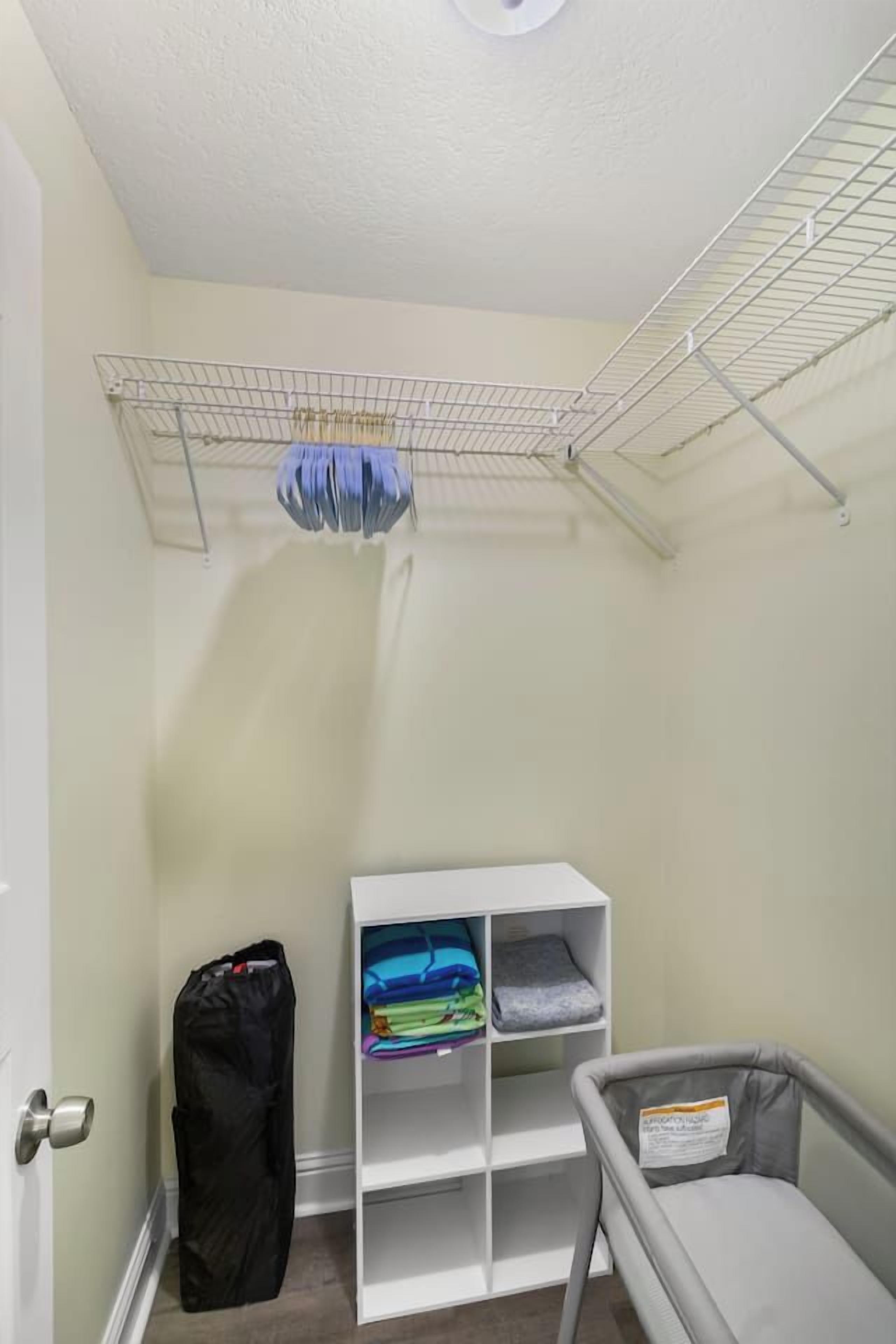 Una habitación con una puerta blanca, un tendedero metálico, un estante blanco con ropa doblada y una cuna infantil gris.