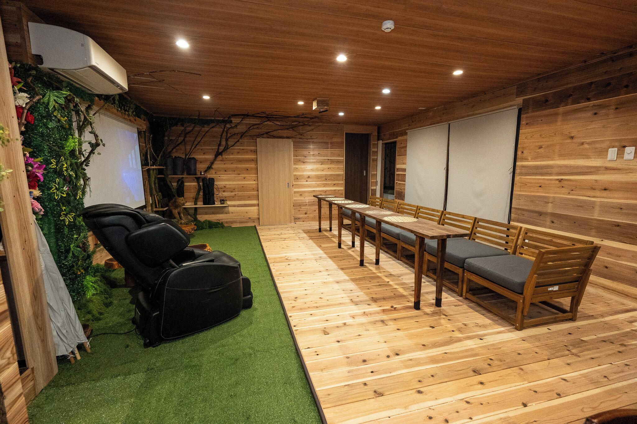A room with a massage chair, a wooden table with chairs, and a green artificial turf area.