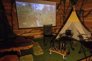 Ein gemütliches Indoor-Camping-Arrangement mit einer Projektionsleinwand, die eine Waldszene zeigt, einem Zelt, Campingstühlen und einem kleinen Tisch mit Tassen und einem Thermos.