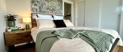 A bedroom with a bed, a nightstand, a lamp, a potted plant, and a wall art piece.