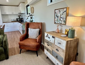 A cozy living room with a brown armchair, a wooden sideboard with drawers, a green vase, and a painting on the wall.