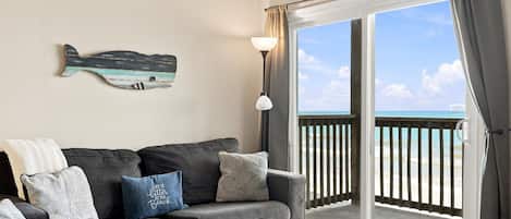 Sala de estar con un sofá gris, cojines estampados, una mesita y vista a la playa a través de un ventanal corredero.