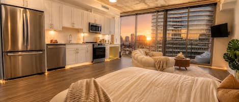 A modern bedroom with a large bed, a city view through floor-to-ceiling windows, and a kitchenette with stainless steel appliances.