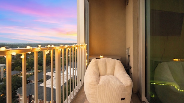 Un balcón con un sillón informal blanco, barandilla con luces de cuerda y vistas a edificios y árboles.
