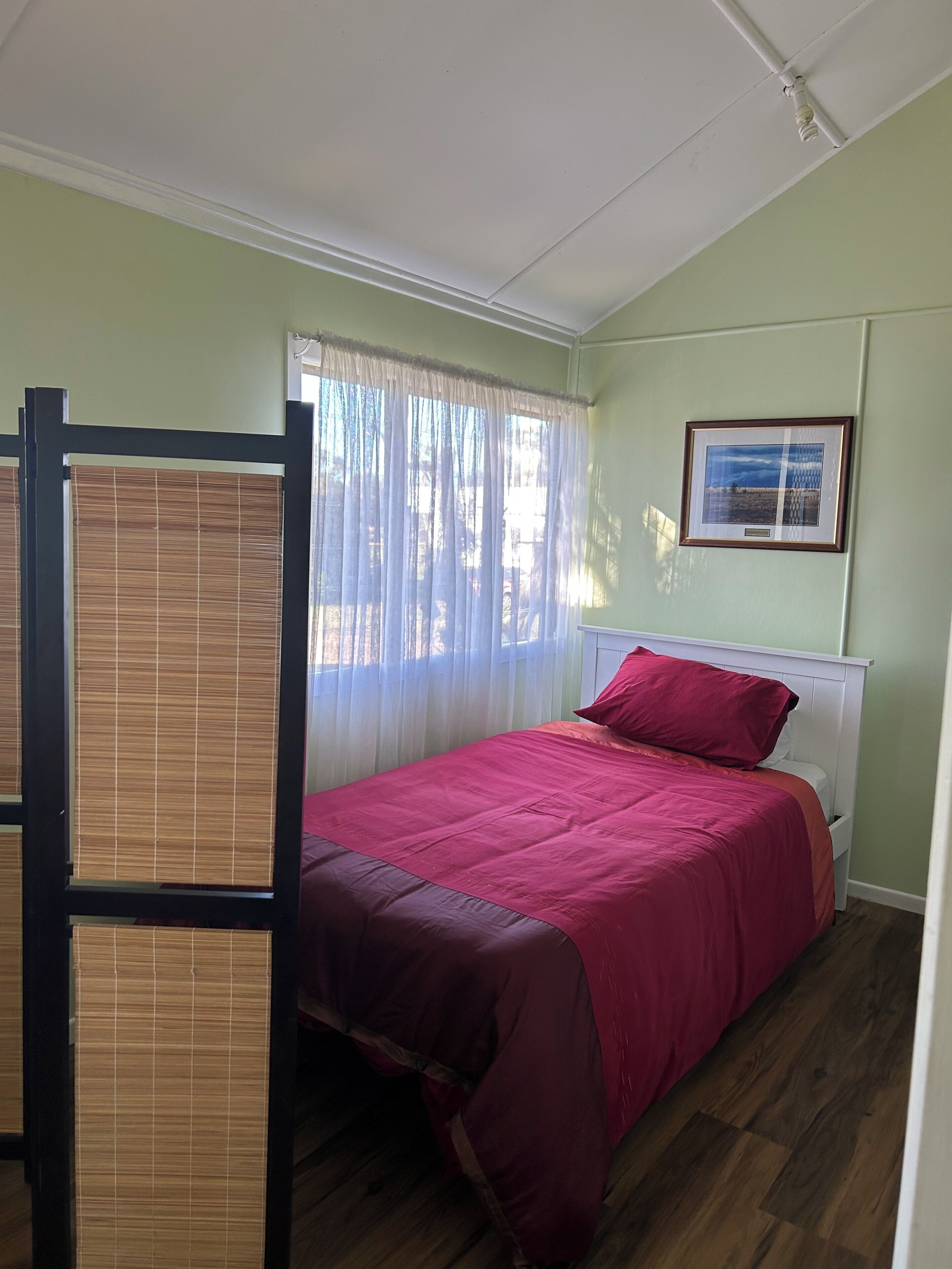 Una habitación con cama, ventana con cortinas traslúcidas, un cuadro en la pared y un ventilador de techo.
