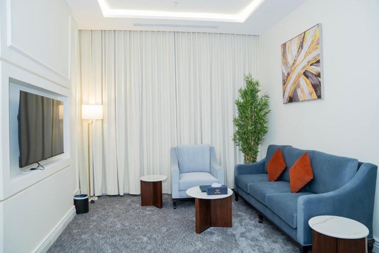 A hotel room with a flat-screen TV, a light blue armchair, a blue sofa with orange cushions, a small round table, and a potted plant.