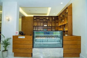 A reception area with a glass display case containing beverages, wooden shelving with various items, and a potted plant. - Brai Hotel (Riyadh)