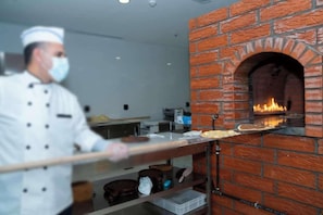 A chef in a white uniform operating a brick oven pizza oven. - Brai Hotel (Riyadh)