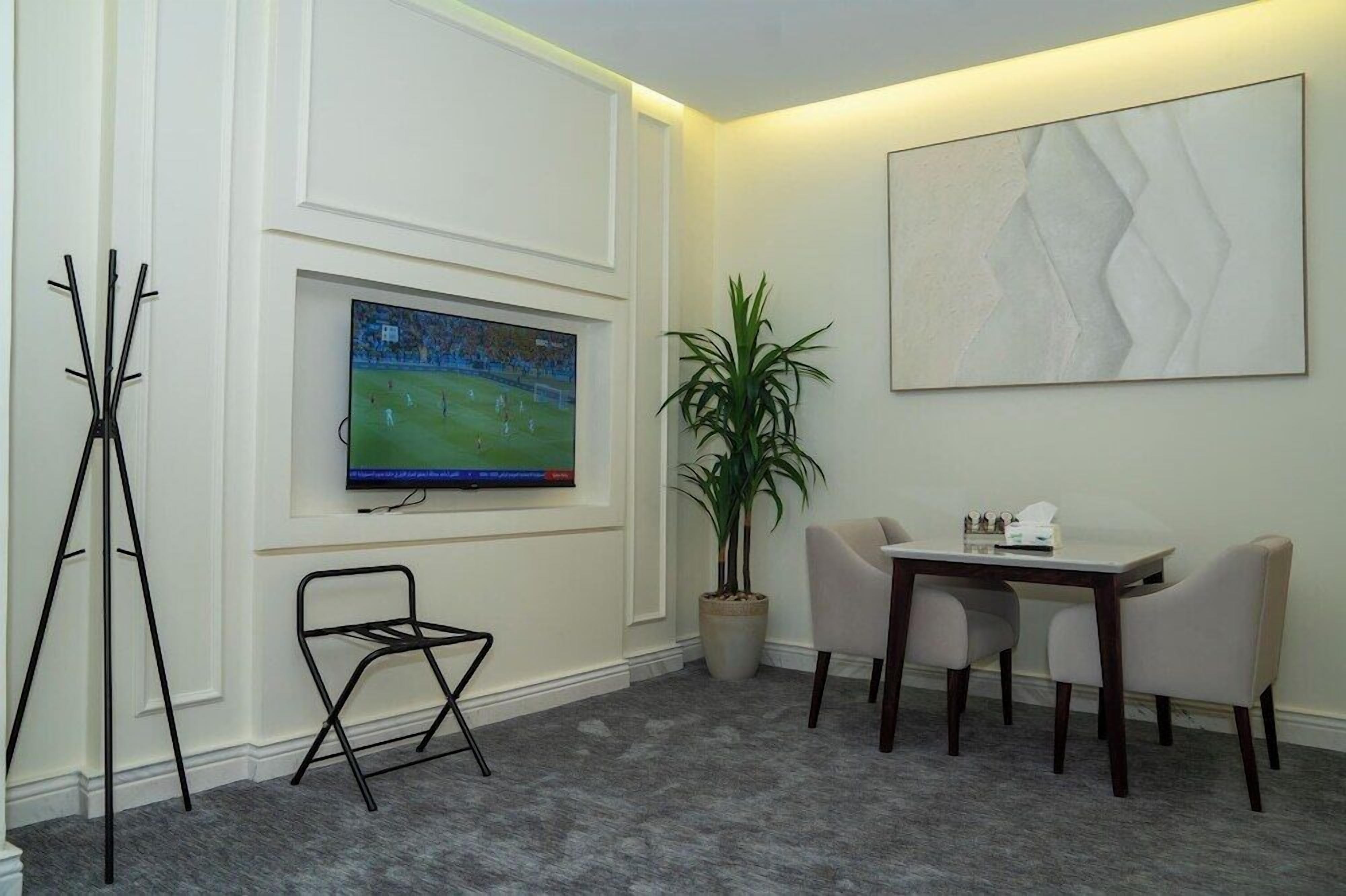 A room with a flat-screen TV mounted on the wall, a potted plant, a small table with chairs, and a stand-alone chair.