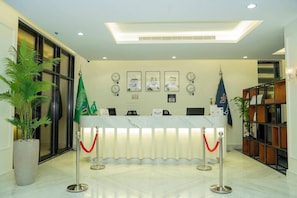 A reception area with a marble counter, flags, framed pictures, and a potted plant. - Brai Hotel (Riyadh)