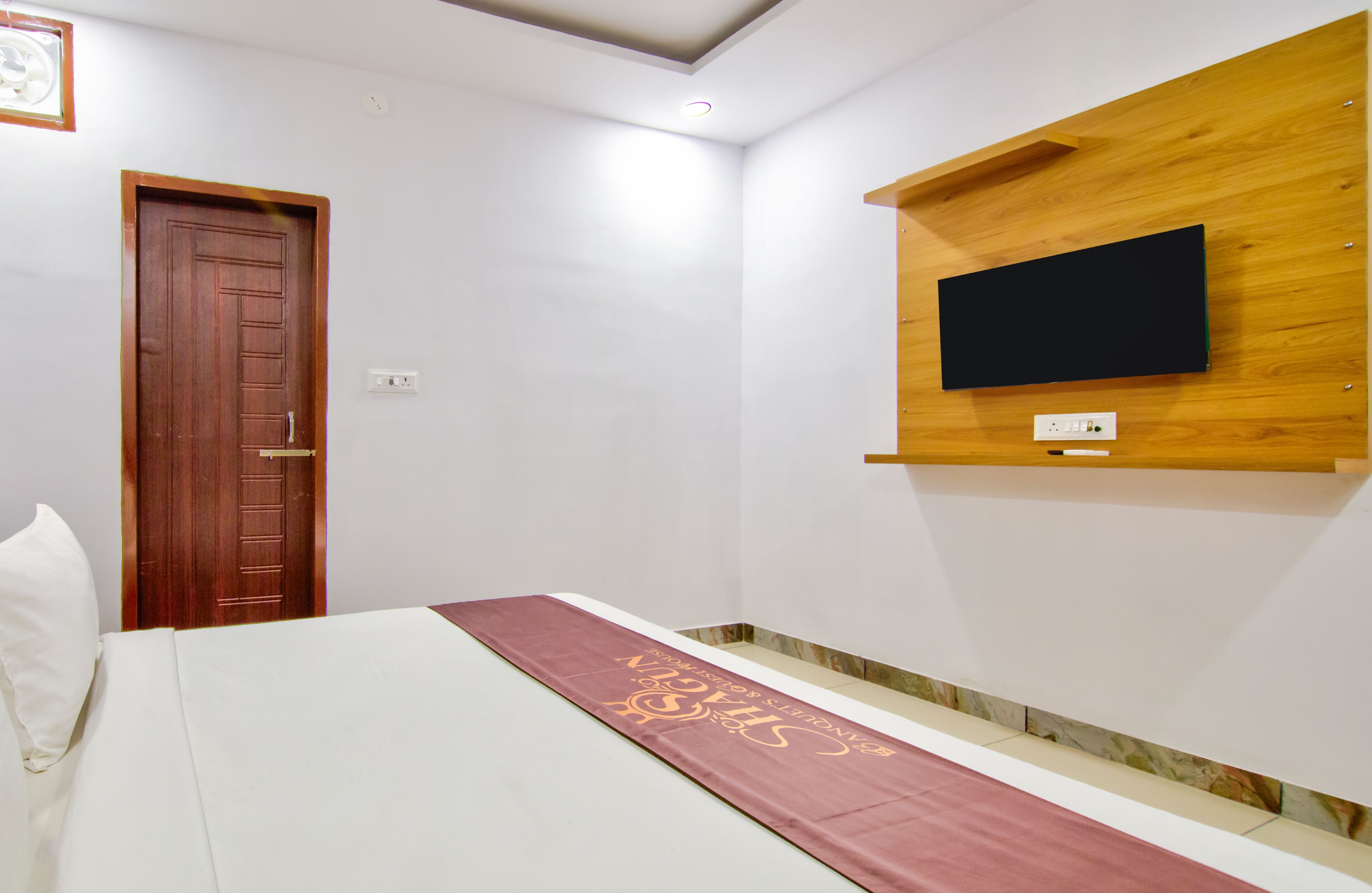 A room with a bed, a wooden wall-mounted TV, a door, and a power outlet.