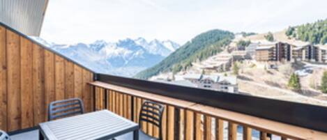 A wooden balcony with a table and chairs, overlooking a mountainous landscape with buildings.