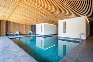 Una piscina interior moderna con techo de madera, agua azul cristalina y una zona de estar con sillas.