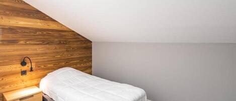 Un dormitorio pequeño y ordenado, con techo inclinado, una cama individual, una mesita de noche y una lámpara fijada a la pared.