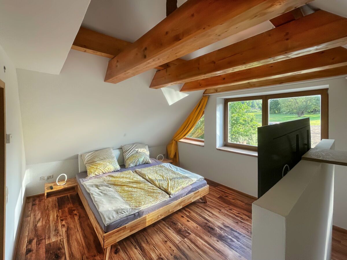 Une chambre avec un plancher en bois, un lit recouvert d’une literie à motifs, une télévision et une fenêtre donnant sur un paysage verdoyant.