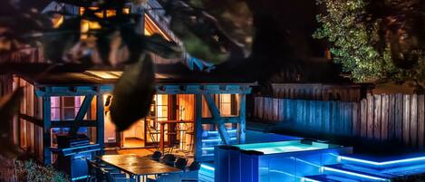 Un espacio exterior bien iluminado con mesa y sillas, una parrilla y una piscina con escalones iluminados.