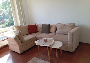 A living room with a beige sectional sofa, a small white table, and a window with blinds. - Charming 2-room vacation apartment with a balcony (Braunlage)