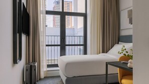 A hotel room with a bed, a desk, a chair, a TV, and a window with a view of buildings.