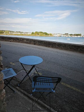 Blick auf das Meer mit einem Tisch und Stuhl auf einer gepflasterten Fläche, mit Blick auf ruhiges Wasser und einen Yachthafen mit Booten.