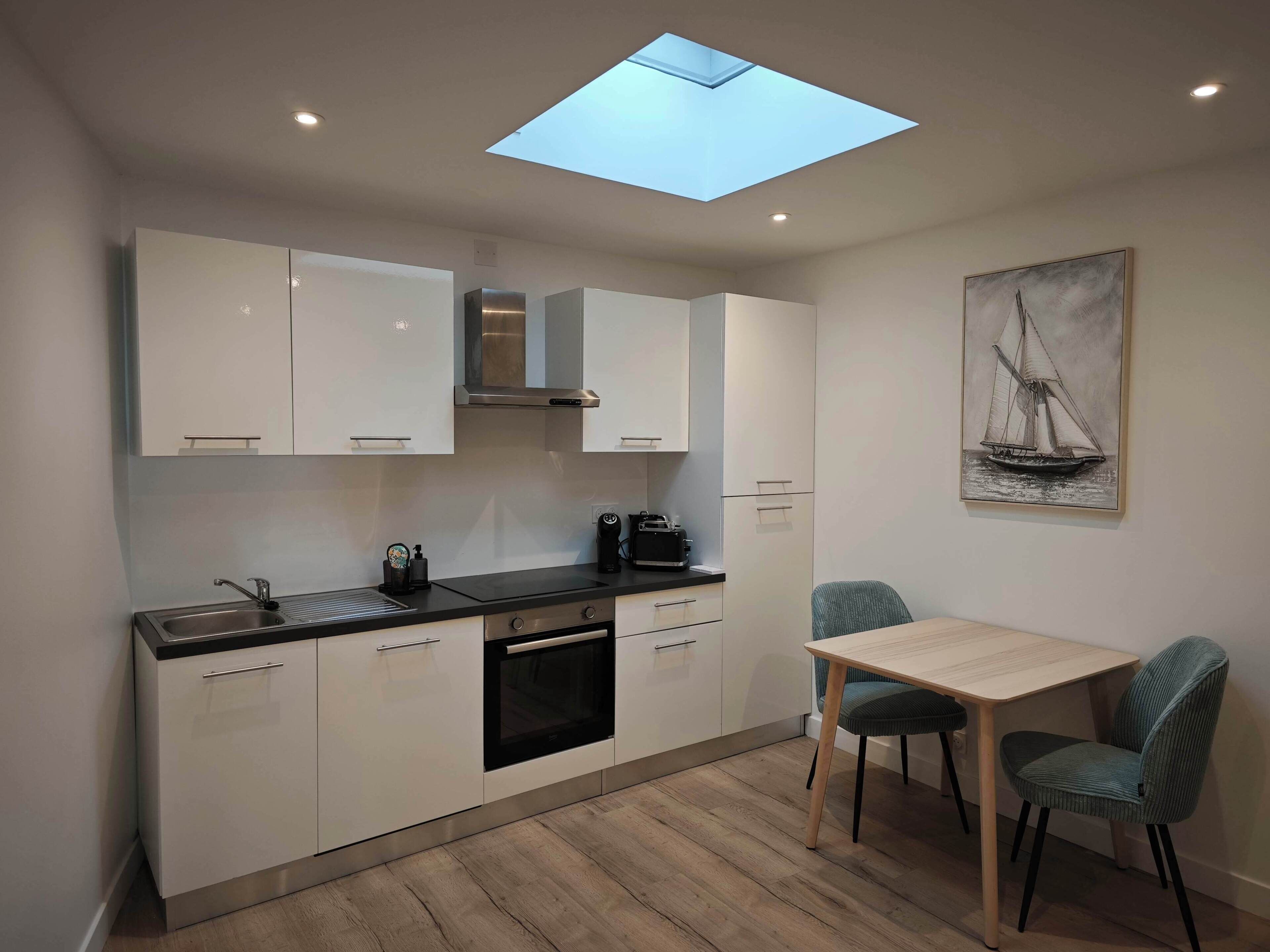 Une cuisine moderne avec des meubles blancs, un puits de lumière, un coin repas avec une table et des chaises, et un tableau accroché au mur.