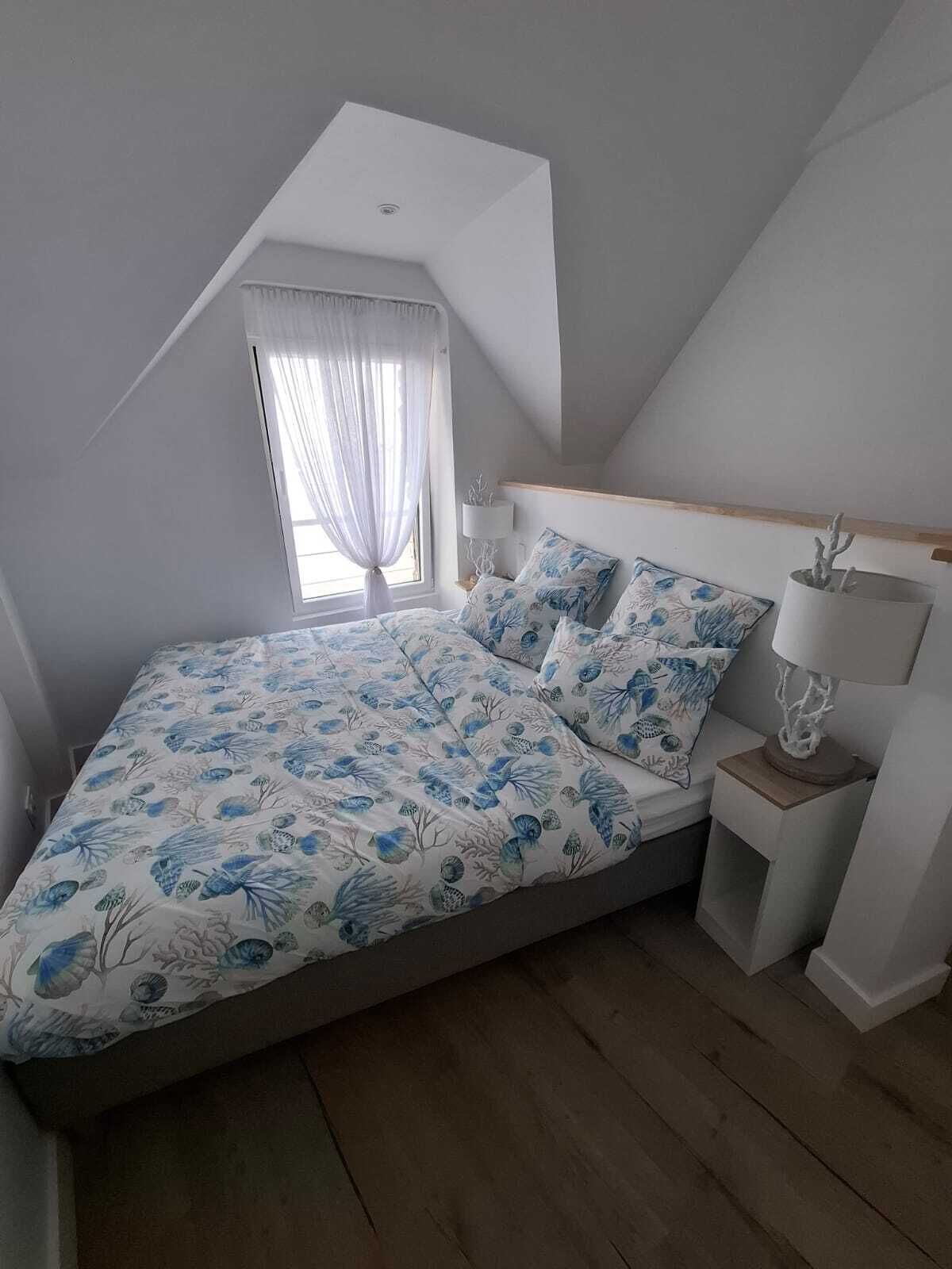 A bedroom with a sloped ceiling, a bed with floral bedding, a bedside table with a lamp, and a window with sheer curtains.