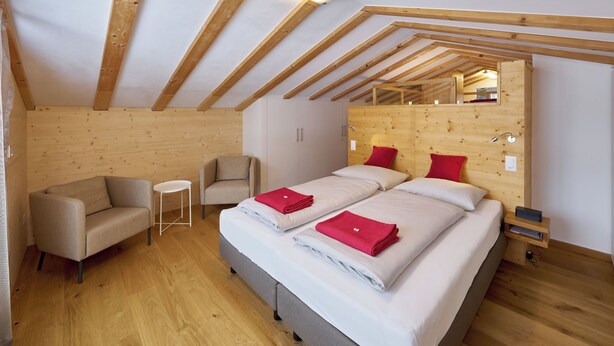 Una camera da letto con soffitto spiovente, travi in legno, un letto con biancheria rossa e bianca, un divano e un piccolo tavolo.