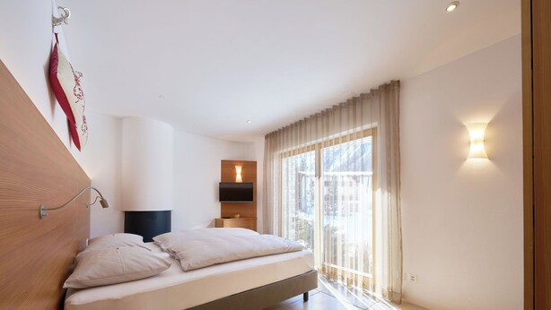 A modern bedroom with a bed, bedside table, and a view of the outdoors through large windows. - Apartments Grischuna Mountain Lodge, (Samnaun-Dorf) by Interhome (Samnaun)