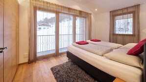 Ein Schlafzimmer mit einem großen Bett, zwei mit Vorhängen versehenen Fenstern, Holzboden und einem Balkon mit Geländer.