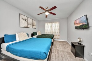 Ein Schlafzimmer mit einem Bett, einem Deckenventilator, einem Fenster mit Vorhängen, einem an der Wand befestigten Fernseher und einem kleinen Nachttisch.