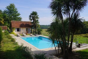 Ein Schwimmbecken mit Liegestühlen, ein Haus mit Ziegeldach und umliegendes Grün.